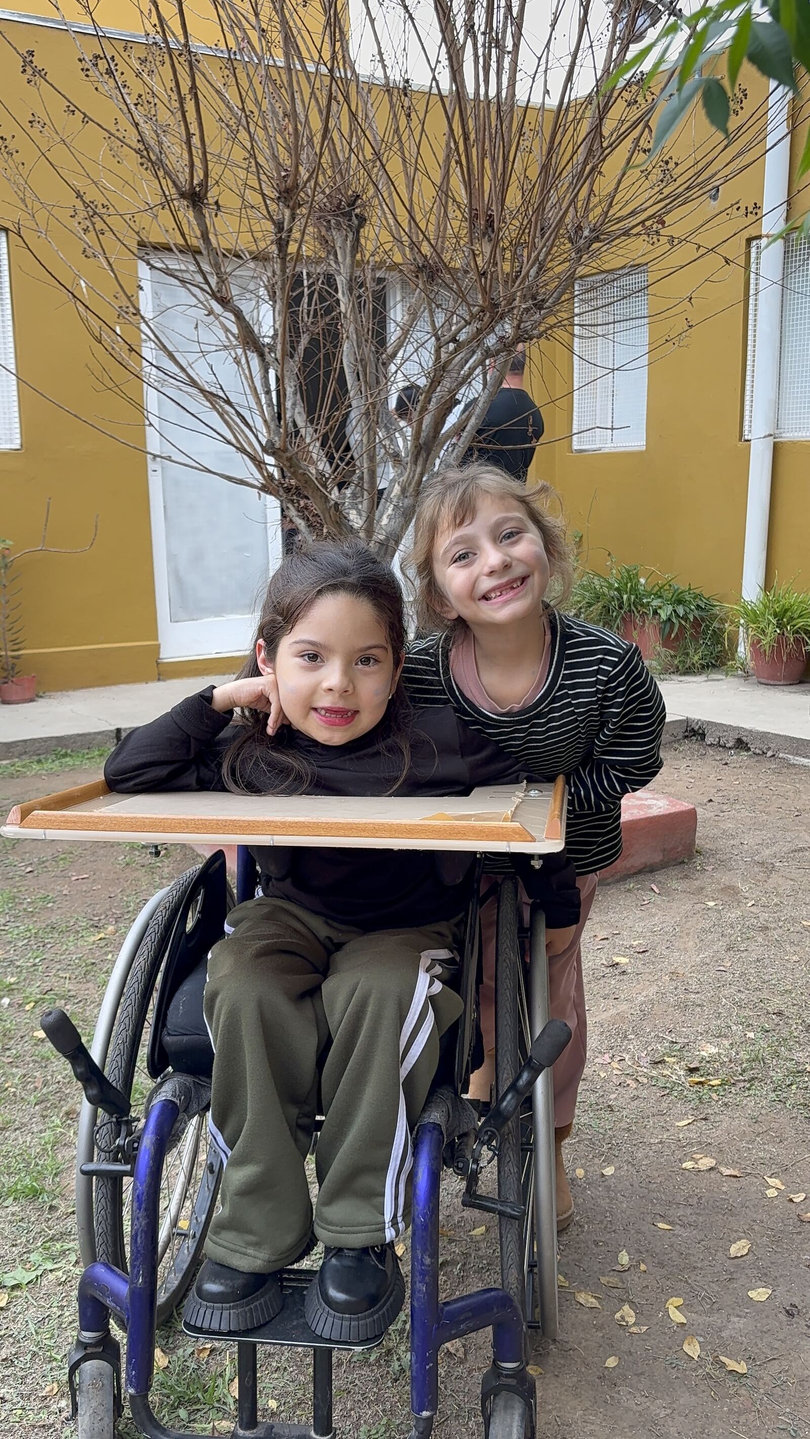 Dos nenas en el patio: una en silla de ruedas, la otra abrazándola por detrás.